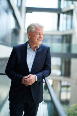 Ein Mann im Anzug steht auf einem Balkon und blickt mit nachdenklichem Gesichtsausdruck in die Ferne. Dieses klassische Geschäftsporträt ist vor der Kulisse eines modernen Gebäudes aus Glas und Stahl gerahmt und fängt seine nachdenkliche Stimmung ein. constantin ranke fotografie