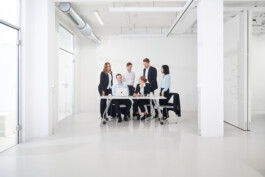 Eine Gruppe von sieben Personen, alles Anwälte und Notare in Geschäftskleidung, versammelt sich in einem hellen, modernen Büro um einen Tisch mit einem Laptop. Sie scheinen in eine Diskussion oder Besprechung vertieft zu sein, mit weißen Wänden und Glastüren im Hintergrund.
