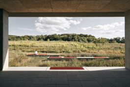 Von der schattigen Betonstruktur aus entfaltet sich eine elegante Darstellung der Architektur. Mit Blick auf ein Grasfeld mit einer entfernten Baumreihe unter einem teilweise bewölkten Himmel genießt eine Person diese Szene von einer modernen Bank aus, die sich nahtlos in die Landschaft einfügt und die Essenz der Fotografie einfängt.