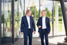 Zwei Männer in Geschäftsanzügen gehen vor einem modernen Glasgebäude und unterhalten sich. Sie scheinen in ein Gespräch vertieft zu sein, im Hintergrund sind Grünflächen zu sehen. constantin ranke fotografie