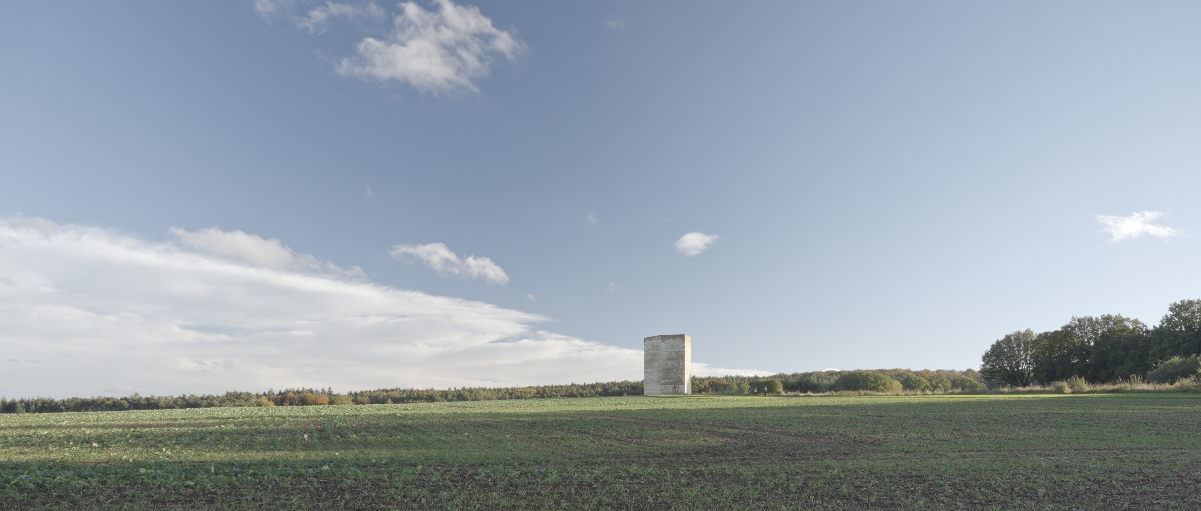 Ein großes, offenes Feld erstreckt sich bis zum Horizont. In der Mitte steht unter einem teilweise bewölkten Himmel eine einsame zylindrische Steinstruktur: die Bruder-Klaus-Feldkapelle von Peter Zumthor. Im Hintergrund sind Bäume zu sehen. constantin ranke fotografie