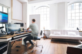 In einem eleganten Ambiente, das sich perfekt für Innenraumfotografie eignet, sitzt eine Person in einem modernen Musikstudio, das mit natürlichem Licht durch große Fenster gefüllt ist. Zwischen Gitarren und moderner Audioausrüstung wird das helle Ambiente durch ein weißes Sofa an der Wand vervollständigt.