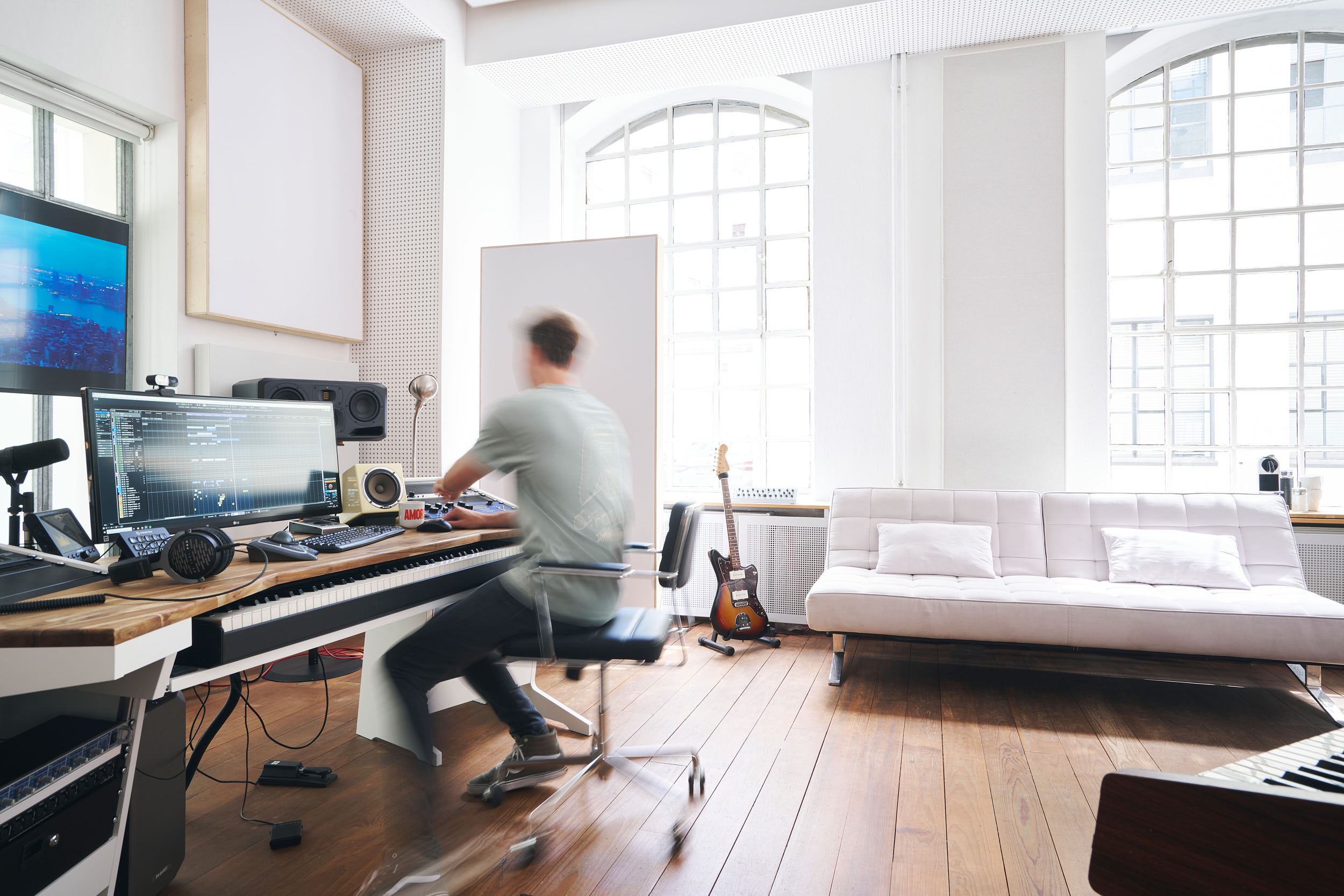 In einem eleganten Ambiente, das sich perfekt für Innenraumfotografie eignet, sitzt eine Person in einem modernen Musikstudio, das mit natürlichem Licht durch große Fenster gefüllt ist. Zwischen Gitarren und moderner Audioausrüstung wird das helle Ambiente durch ein weißes Sofa an der Wand vervollständigt. constantin ranke fotografie