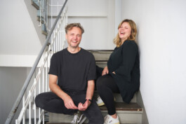 Ein Mann und eine Frau sitzen auf einer Innentreppe und lächeln beide in einem entspannten Moment, der durch Innenraumfotografie festgehalten wurde. Der Mann, in ein schwarzes Hemd und eine schwarze Hose gekleidet, sitzt auf einer unteren Stufe, während die Frau in einem schwarzen Kleid und Leggings auf einer höheren Stufe an der Wand lehnt.