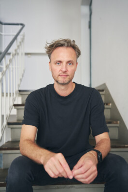 Ein lächelnder Mann mit kurzen Haaren und Bart, der ein schwarzes T-Shirt trägt, sitzt lässig auf einer Treppe. Die Kulisse verkörpert mit ihrem Geländer und dem schlichten Wandhintergrund die Essenz der Innenraumfotografie und schafft eine entspannte und ungezwungene Atmosphäre.