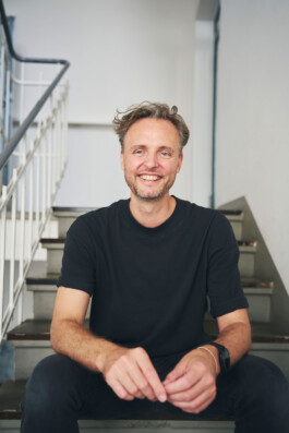 Ein lächelnder Mann mit kurzen Haaren und Bart, der ein schwarzes T-Shirt trägt, sitzt lässig auf einer Treppe. Die Kulisse verkörpert mit ihrem Geländer und dem schlichten Wandhintergrund die Essenz der Innenraumfotografie und schafft eine entspannte und ungezwungene Atmosphäre.