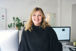 Eine Person mit schulterlangem blondem Haar und schwarzem Pullover lächelt in einem modernen Büro. Ein Computerbildschirm und eine Topfpflanze fangen die Essenz der Innenraumfotografie ein und verbessern subtil die Atmosphäre im Hintergrund.