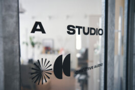 Glastür mit dem Text „A Studio“ und „Creative Audio“ neben abstrakten Formen. Die unscharfe Innenraumfotografie lässt Andeutungen von Grünpflanzen und stilvollen Möbeln im Hintergrund erkennen.